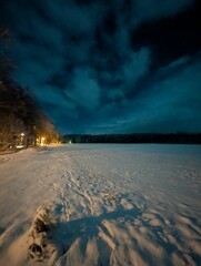 A tranquil night scene featuring a snowy landscape and a dramatic sky filled with dark clouds. The ground is covered in a thick layer of snow, with soft moonlight