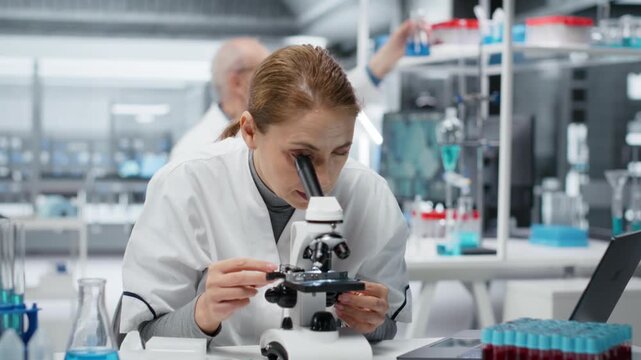 Female microscopist examining microscope glass slide in laboratory to view cells. Research facility employee checks sample slide, doing imaging process using optical device, camera B