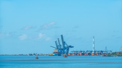 Commercial cargo port with giant blue cranes, container ships, and industrial chimneys under a bright blue sky. Global logistics and shipping industry