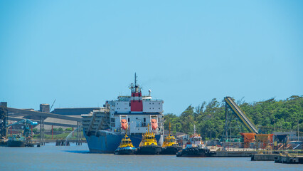 Massive industrial cargo ship docked at a port, flanked by four colorful tugboats providing support. The scene captures heavy maritime infrastructure, including loading conveyors and harbor cranes