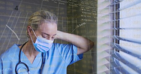 Standing nurse rubbing neck at clinic window with blinds, in blue scrubs, mask, stethoscope, charts