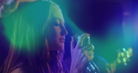 Singing female singer in dark outfit holding retro mic on stage with colored lights, blurred guitar