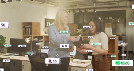 Discussing two coworkers in blouses reviewing tablet and papers at office desk with social overlays