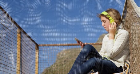 Sitting child in sweater touching green headset, holding phone on deck by mesh fence, copy space