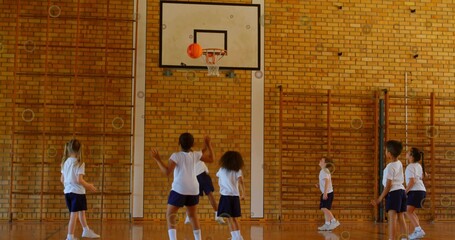 Shooting seven kids in white tees and navy shorts practicing under gym hoop with orange basketball