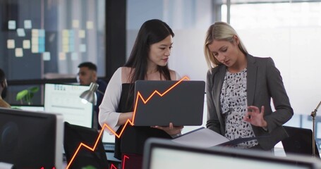 Showing laptop and flipping binder, two women in blouses discussing at office, orange upward graph