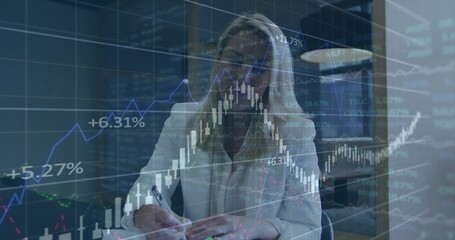 Writing mature analyst in white blazer and glasses in office using pen and notes, charts overlaying