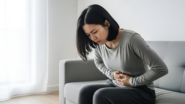 Young asian woman experiencing acute abdominal pain while sitting on a sofa at home