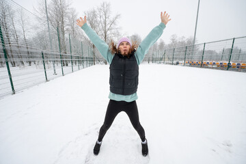 Girl does an exercise where she jumps in place with her arms up and her legs apart on a snowy sports field in winter.