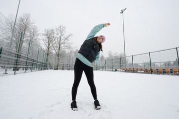 Girl doing a side bend exercise with her arm raised on a snowy sports field in winter.