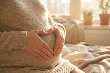 Pregnant woman gently cradling her baby bump with hands forming a heart shape, bathed in warm, soft sunlight indoors.