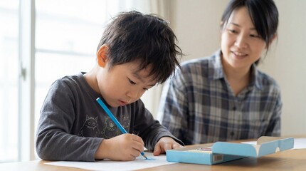 Focused asian boy drawing with blue pencil on paper while smiling mother watches him at home table