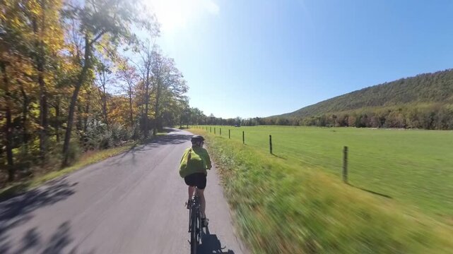 A mature woman rides her bike on a road beside the river in West Virginia surrounded by autumn trees and bright sunshine.