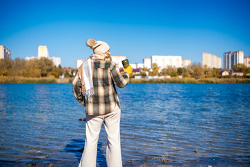 Woman holding travel mug standing by lake facing city in autumn. Quiet outdoor pause supporting...