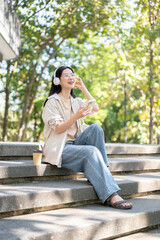 Obraz premium Glasses asian woman wearing headphones listening to music while sitting on concrete stairs in park.