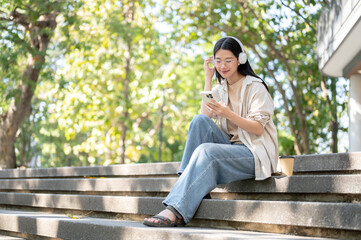 Obraz premium Glasses asian woman holding headphones listening to music while sitting on concrete stairs in park.