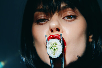 Stylish woman with red lips holding sushi with chopsticks, showcasing bold fashion and elegance in a modern culinary setting.