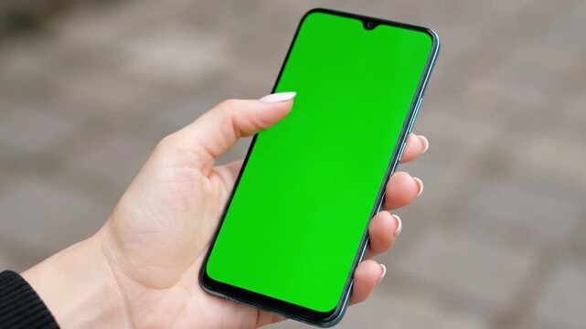 A person's left hand is shown holding a smartphone with a green screen for easy customization outdoors.