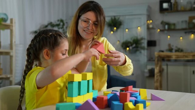 Building colorful block tower together mother and daughter concentrate to stack block and toy on table in cozy kitchen while child learn through play creating family bond and playful learning