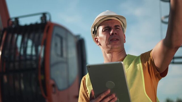 Worker inspects tablet on construction site wearing hard hat and vest near crane and heavy machinery while engineer reviews digital plan for safety inspection and equipment checklist and records data - Powered by Adobe