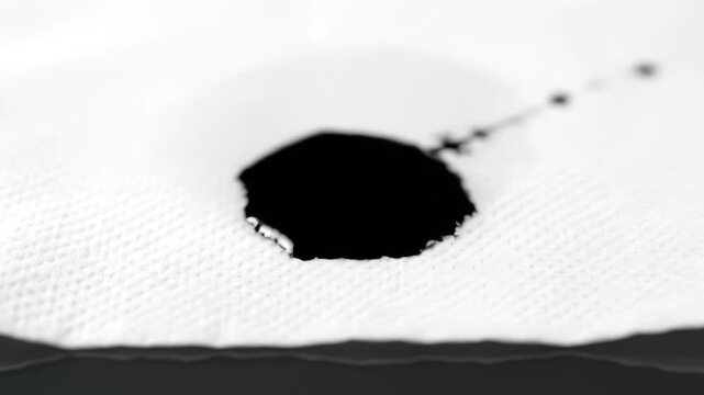 A dramatic slow-motion macro shot of a single black ink drop falling onto white absorbent paper, showing the liquid spreading and seeping into the textured fibers