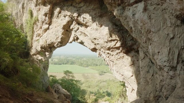 Cinematic video panning through the natural rock arch of Khao Talu Miti. The cave frame reveals a lush green landscape in Ratchaburi.