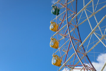 Colorful Ferris wheel gondolas against bright blue sky and clouds.