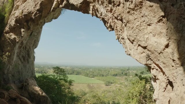 Cinematic video panning through the natural rock arch of Khao Talu Miti. The cave frame reveals a lush green landscape in Ratchaburi.