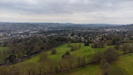 Obraz premium View across a park in Bath, UK, with bare trees, bordering roads and houses, and the city extending into rolling hills and countryside beneath an overcast sky.