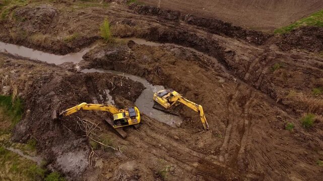 Two yellow excavators work in unison to clear debris and reshape the muddy terrain, creating a water channel in a construction site with visible earth and machinery