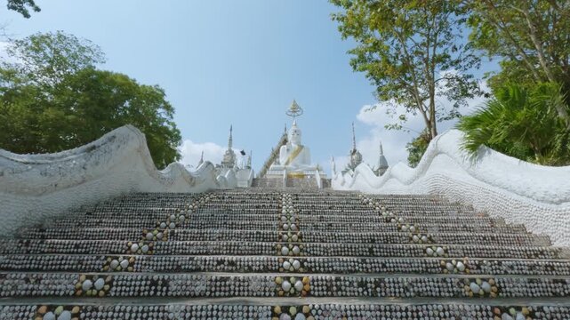 Wide shot of the magnificent white chapel at Wat Phu Nam Ron, Ratchaburi. The ornate Thai temple stands beautifully under a bright blue sky with clouds.