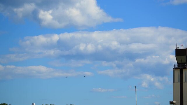 Two military fighter jets fly past an airport control tower against a blue sky with drifting clouds, creating a dynamic aviation scene with airbase infrastructure.