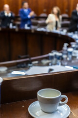 An empty coffee cup on a wooden desk with the blurred background of a plenary session in a...