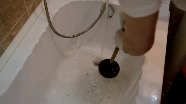 Individual using a black plunger to clear a clogged bathtub drain, demonstrating the action of plunging water and debris in a bathroom setting with visible plumbing tools