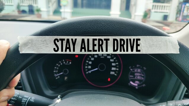 Hands on the steering wheel of a car and a sticker with the words Stay alert drive. concept of driving cautiously or carefully on the road - Powered by Adobe