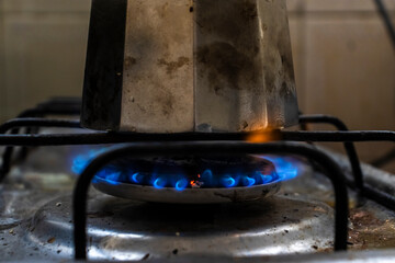 A classic Italian moka pot sits on a lit gas stove. The bright blue flames heat the base, illustrating the traditional coffee brewing process in a domestic kitchen setting.