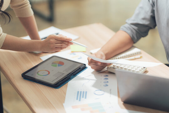 Business Meeting Discussion: Close-up of hands-on data analysis, two business professionals are huddled over a desk, engrossed in a discussion about charts and graphs on papers and a tablet.