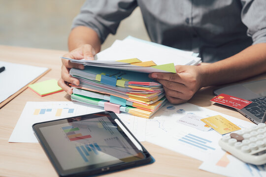 Overwhelmed with paperwork: A focused professional meticulously manages a towering stack of files, navigating a workspace brimming with financial documents and digital tools. - Powered by Adobe