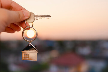 Hand holding house key with home keychain in front of blurred city at sunset, symbolizing real...