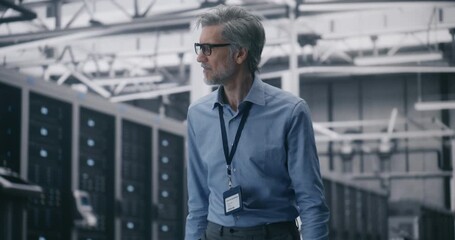 Experienced Engineer Overseeing Cloud System Integrity. Mature IT Specialist With Glasses Walks and Monitors Systems Clean, Bright Server Hall. Concept of Mobile System Management and Data Integrity.