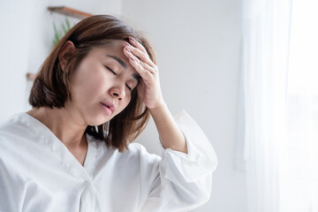 Asian woman sitting on bed in morning, touching head with hands, suffering from painful headache, migraine, or vertigo. Sick female feeling dizzy after waking up in bedroom