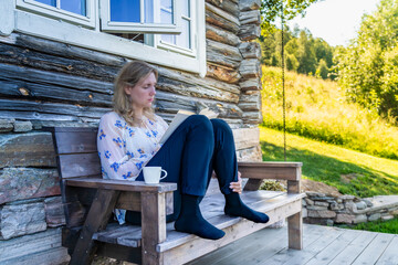 A woman sits on a wooden bench in front of a log cabin engrossed in a book. Enjoying her me time reading a book with a cup of tea in the Shade on a Sunny Day