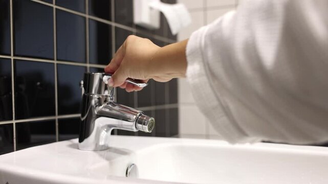 A woman's hand turns on the water bidet with a chrome faucet in a tiled bathroom. The wall has a mix of dark and light tiles, creating a stylish contrast.