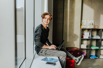Student sitting on windowsill with laptop in classroom smiling