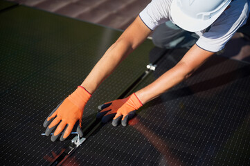 Worker building photovoltaic solar panel system on rooftop of house. Close up of man engineer in helmets and gloves installing solar module with help of hex key outdoors. Renewable energy.