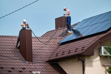 Workers installing solar panels system on rooftop of house. Electricians connecting cables from solar modules to inverter through the chimney for generating electricity through photovoltaic effect.