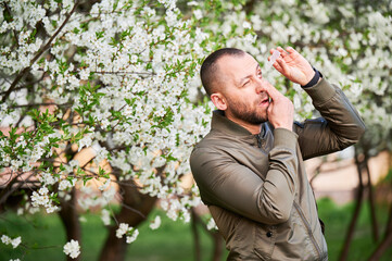 Man allergic using medical eyes drops, suffering from seasonal allergy at spring in blossoming garden. Handsome man treating eyes in front of blooming tree outdoors. Spring allergy concept.