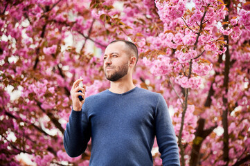 Man allergic using medical nasal drops, suffering from seasonal allergy at spring in blossoming garden. Handsome man showing nasal spray near blooming tree outdoors. Spring allergy concept.