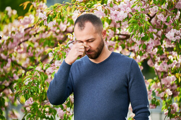 Man allergic suffering from seasonal allergy at spring in blossoming garden at springtime. Bearded young man sneezing and having runny nose in front of blooming tree. Spring allergy concept.