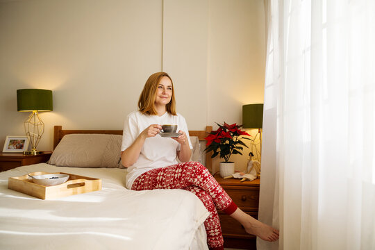 Woman in pyjamas enjoying hot drink on bed with poinsettia indoors